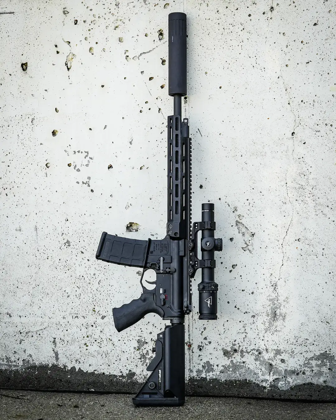 Black AR15 rifle with mounted scope and suppressor leaned against a concrete wall